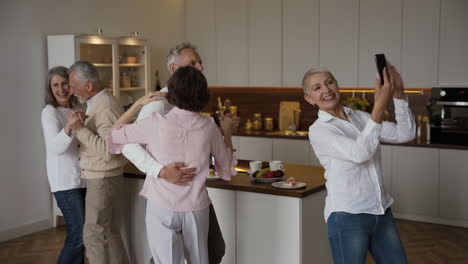 two happy senior couples dancing in the kitchen, while a pretty elderly woman taking a selfie video on mobile phone