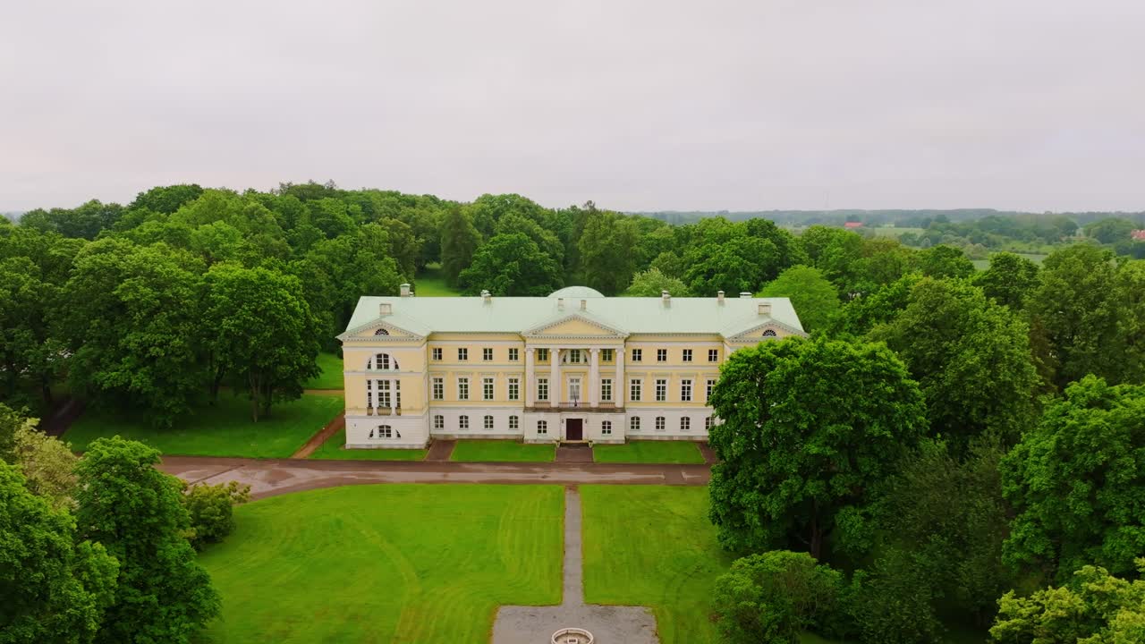 Drone slow recedes from Mežotne Palace framed by green trees under cloudy skies