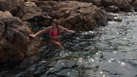 lady in red bikini climbs down into water from stone beach