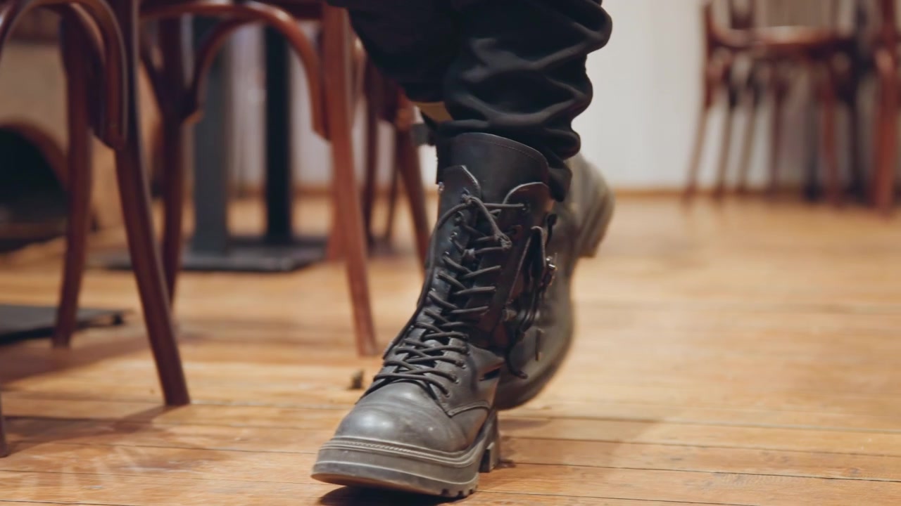 Sepatu bot hitam usang melintasi lantai kayu, tampilan dekat sepatu bot kulit bertali yang lecet melangkah melalui interior bistro yang nyaman dengan kursi kayu bengkok, lantai kayu hangat, kaca reflektif, dan bayangan murung, musisi.