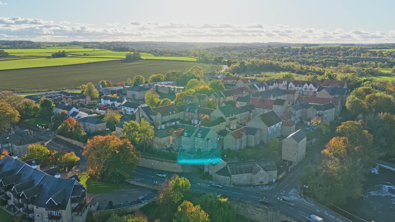 Drone establishing shot showing stone rooftops, residential area, autumn trees and surrounding countryside fields in Wetherby, Yorkshire. Golden hour sunlight, open rural landscape, tranquil mood