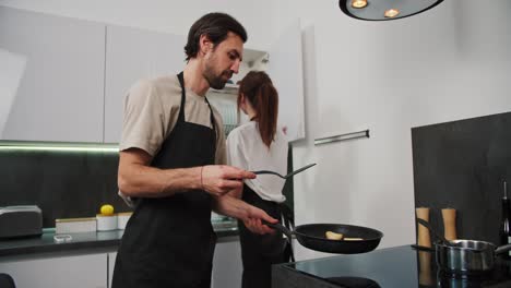 Un-Hombre-Moreno-Feliz-Con-Barba-Incipiente,-Con-Un-Delantal-Negro-Y-Una-Camiseta-Beige,-Prepara-Tartas-De-Queso-Durante-El-Desayuno-Con-Su-Esposa-Morena-En-Un-Apartamento-Moderno.-Cocina-Panqueques-Y-Prepara-El-Desayuno-En-Una-Cocina-Moderna.