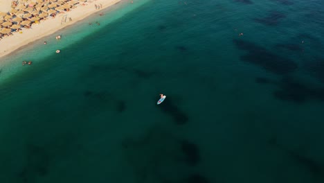 un paddleboarder flota en un mar azul claro cerca de una playa ocupada con sillas de sol y sombrillas