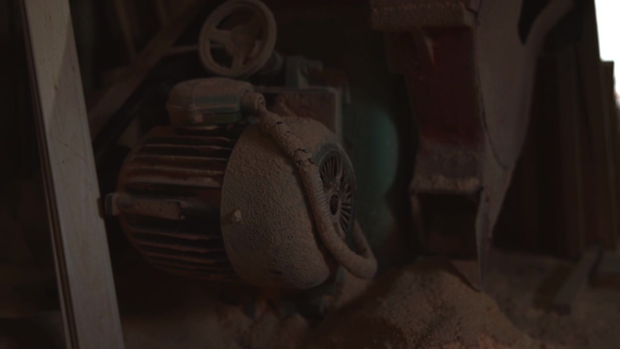 Close-up view of stationary electric motor beside rusted machine parts inside dim workshop, showing finned housing, round fan cover, braided conduit, heavy dust, sawdust, corrosion, and worn metal