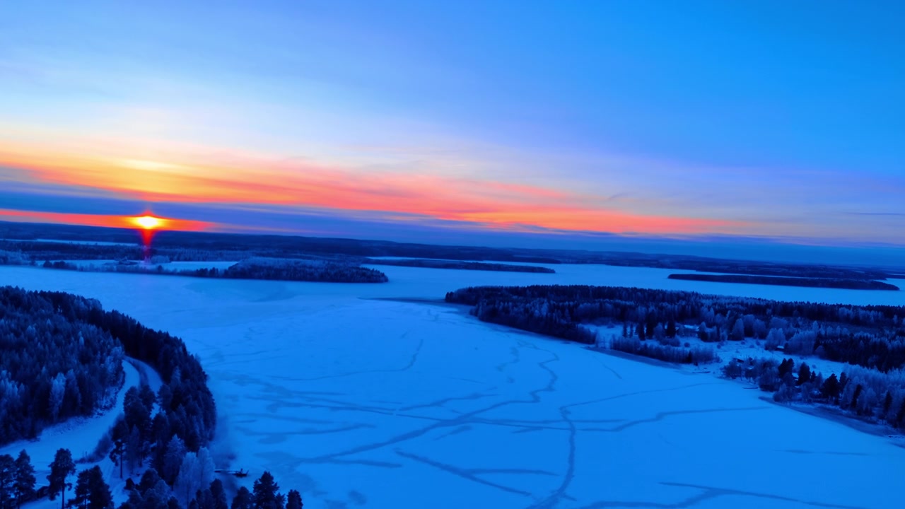 Frossen innsjø ved solnedgang med skogkledde øyer, Lappland, Finland. Droneutsikt over stort isdekket vann med sledespor, skogkledde halvøyer, rød sol i horisonten under oransje og blå himmel