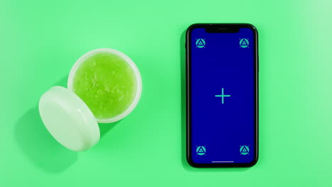 phone with green screen chroma key with fresh ripe avocados with cream and essential oil on green background close up, top view with mint mentol, make up cosmetics