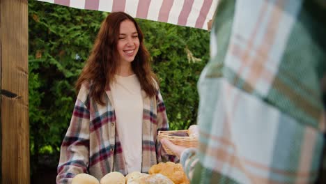 Over-the-shoulder,-a-happy-guy-in-a-green-checkered-shirt-buys-pastries-from-a-brunette-girl-in-a-checkered-shirt-in-a-shop-during-a-fair-near-the-city-against-the-backdrop-of-green-coniferous-trees-in-summer