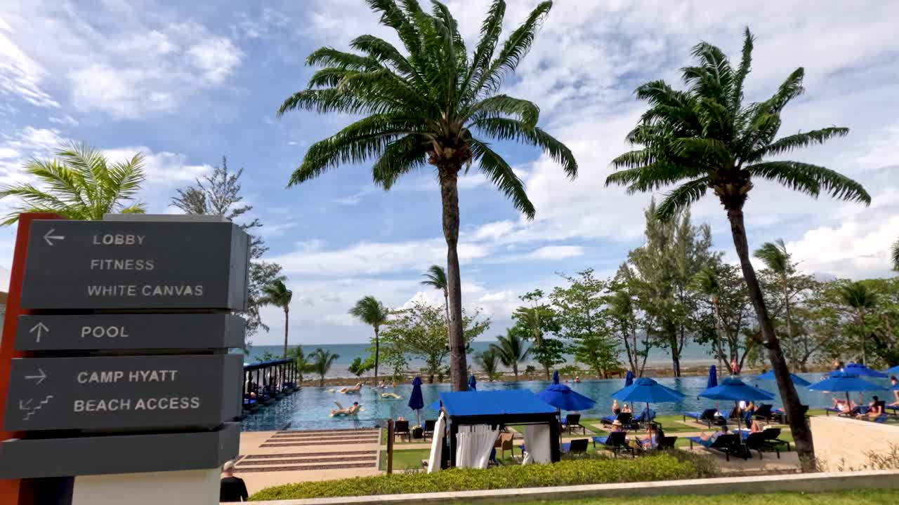 Static shot of resort sign, palm trees, pool, and guests under bright tropical daylight