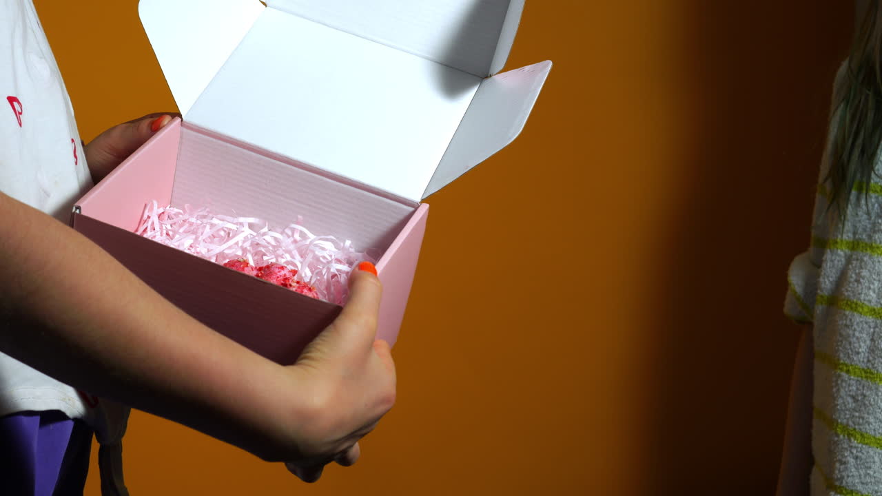 Two children exchange a pastel pink gift box with heart-shaped flaps against a warm orange background, capturing a playful and joyful birthday or celebration moment with hands in focus.