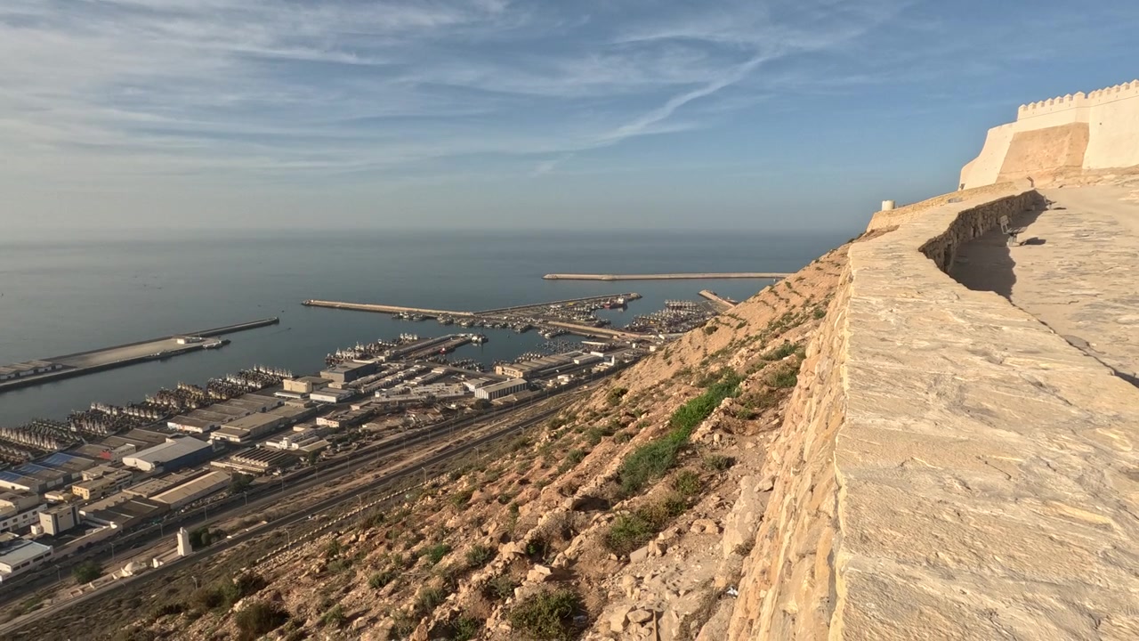 Fortificazione storica in pietra che domina un trafficato porto peschereccio e commerciale, con barche, banchine, moli, acqua blu calma, colline rocciose, vegetazione secca e strutture marittime lungo la costa.