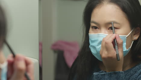 young asian american woman with face mask applying eyebrow makeup