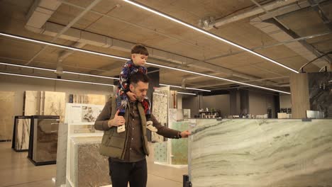 A Young Father with His Son on His Shoulders Walks in a Hardware Store and Examines the Goods. A young father with his son on his shoulders walks in a hardware store and examines the goods. Dad and son in the salon of ceramic tiles