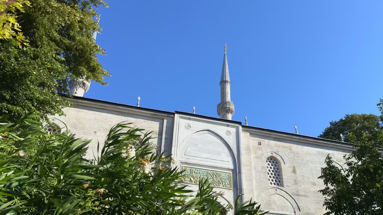 Green trees and sunlit mosque entrance under clear blue sky in summer light, establishing tracking right