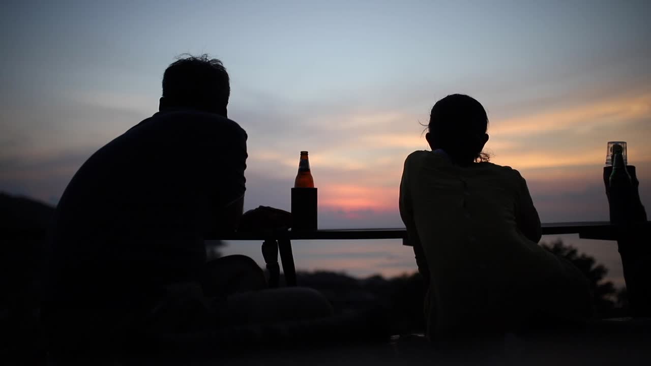 Two silhouetted people watching a beautiful sunset over the ocean whilst having a beer in Thailand.
