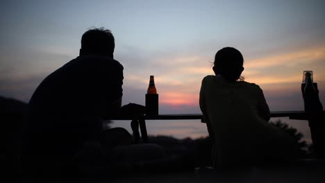 two silhouetted people watching a beautiful sunset over the ocean whilst having a beer in thailand.