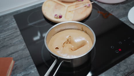 person stirring swollen cornflakes mixture in pot on electric stove, thick, bubbly texture forming as heat causes expansion, kitchen scene with partial view of wooden tray and floral decor