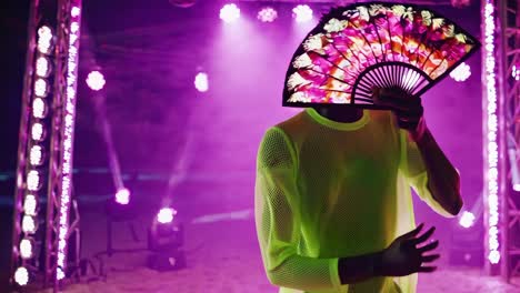 dj hiding face behind a hand fan while performing on stage during a vibrant night beach party, surrounded by purple lights and swirling fog in lively ibiza