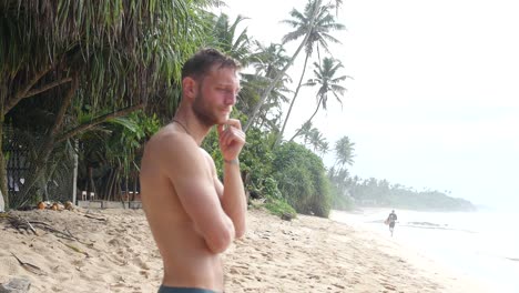 Attraktiver-Mann-Ohne-Hemd-Steht-Am-Tropischen-Sandstrand-Und-Blickt-Auf-Das-Wasser,-Um-Die-Surfbedingungen-Zu-Beurteilen