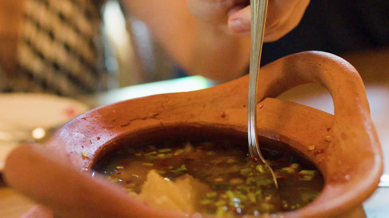Hand lifts spoonful of herbal Thai soup from clay pot, warm ambient lighting, close-up view