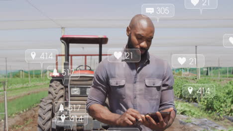 mid-adult farmer tapping smartphone by red tractor on farm with social icons hovering over crops