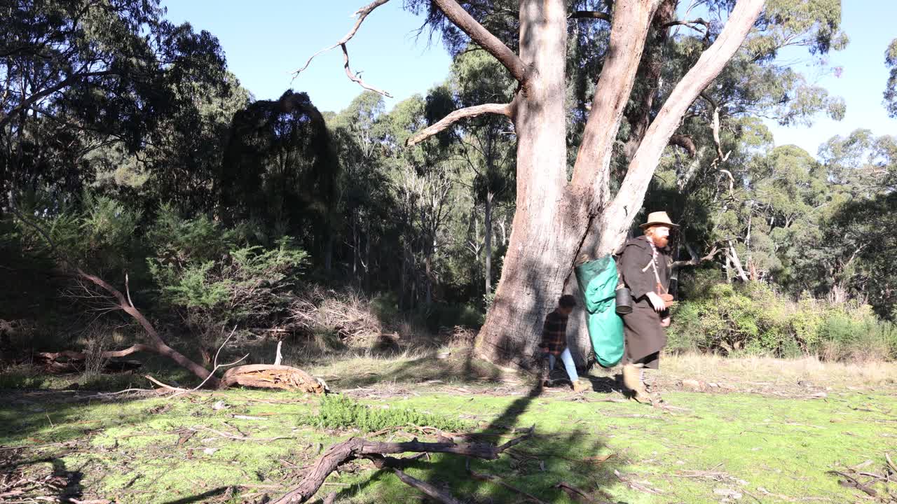A swagman in a stockman jacket and akubra hat walks through the Australian bush with his son.