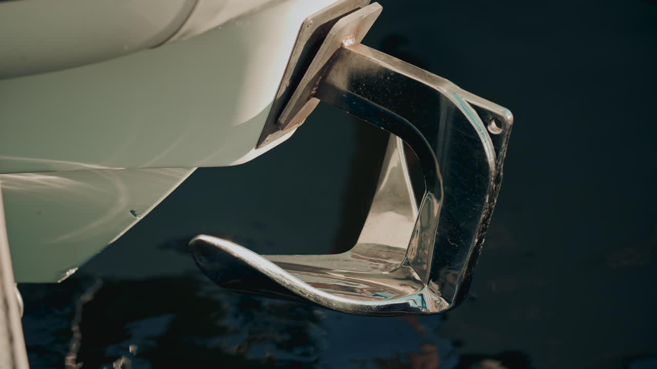 A chrome boat anchor with smooth curves, set against abstract blue and turquoise water reflections in a marina