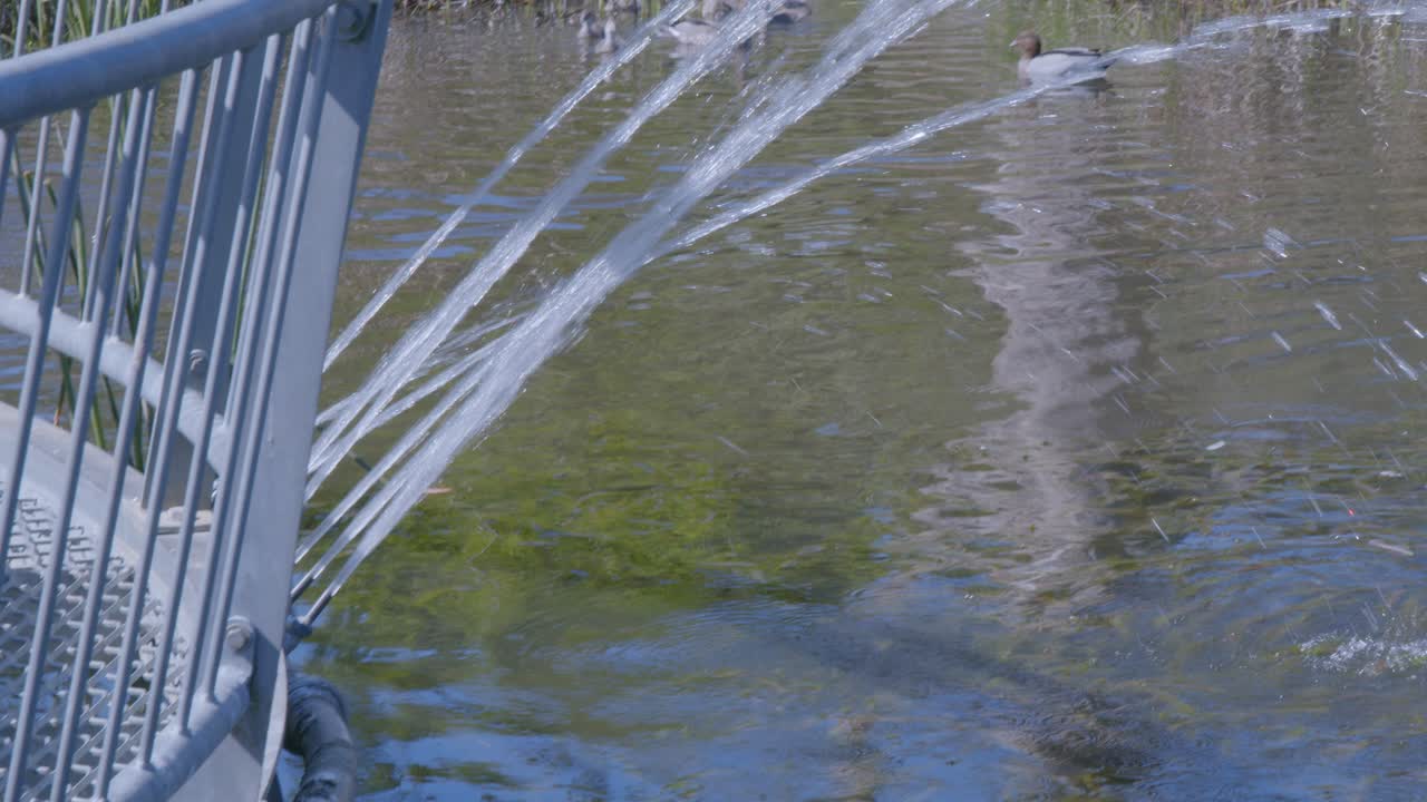 Serene close-up of a metal bridge railing with flowing water beneath, capturing reflections and gentle ripples. Sunlight enhances the peaceful, natural setting
