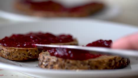 male hand spreading sweet red strawberry jam on top of golden brown toasted flax seed bread with silver knife