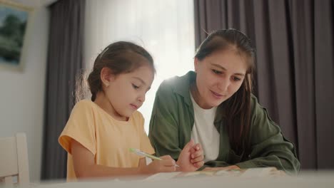 A-little-brunette-girl-in-a-yellow-dress-does-her-homework-with-her-mother,-a-brunette-woman-in-a-green-jacket,-and-writes-in-her-notebook-while-sitting-at-the-table-in-a-cozy-modern-apartment