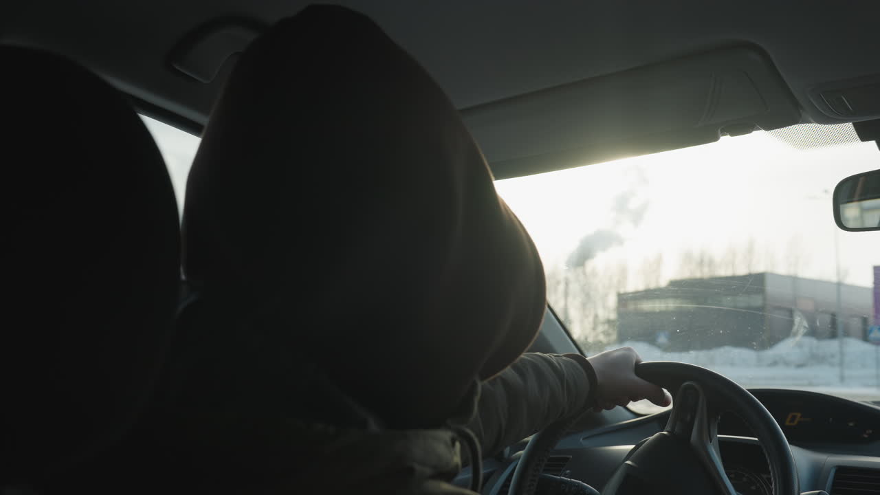 person in hoodie with hood up and tinted glasses gently tapping steering wheel while swaying to music inside sunlit car cabin overlooking snowy urban parking lot and distant building