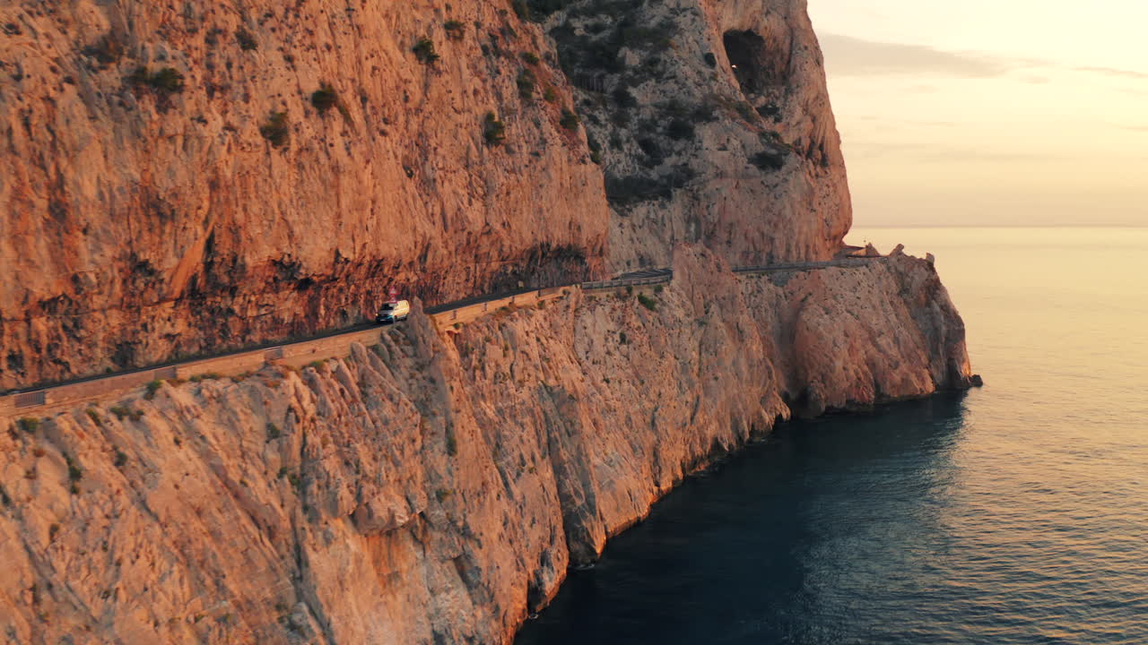 Drone shot of small van driving on otherworldly coastal road winding along steep rugged cliffs bathed in sunrise hues. Golden light reflecting on the sea below. Car on engineering marvel