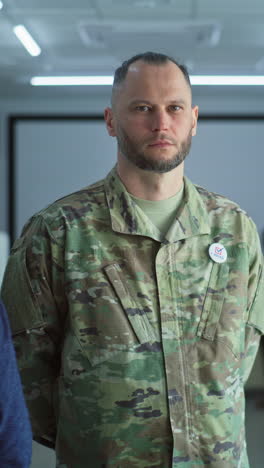 woman in camouflage uniform stands in polling station and looks at camera. portrait of female soldier, united states of america elections voter. background with voting booths. concept of civic duty.