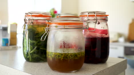 handheld shot of fermented wild garlic, fermented red berries, pickled wild garlic flowers and raspberry vinegar in jars in a kitchen with the chef working in the kitchen in the blurred background