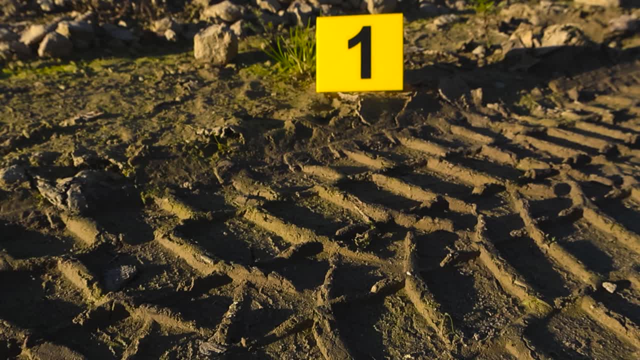Close up view gliding over muddy vehicle or car tire tracks in muddy damp countryside ground with a yellow crime marker next to it, showing it as forensic evicende at a crime scene