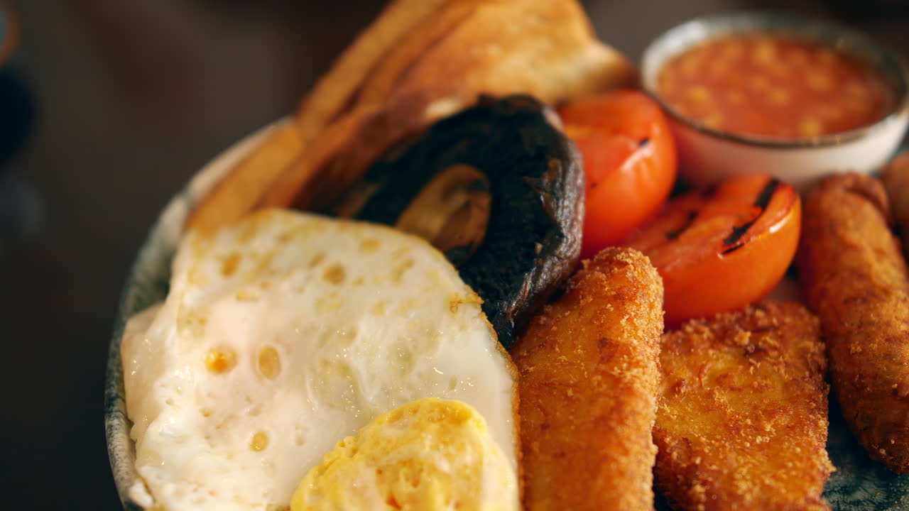 Un piatto di colazione inglese con uovo fritto, pomodoro grigliato, hash brown, salsiccia, toast, funghi e fagioli al forno serviti in una ciotola a parte