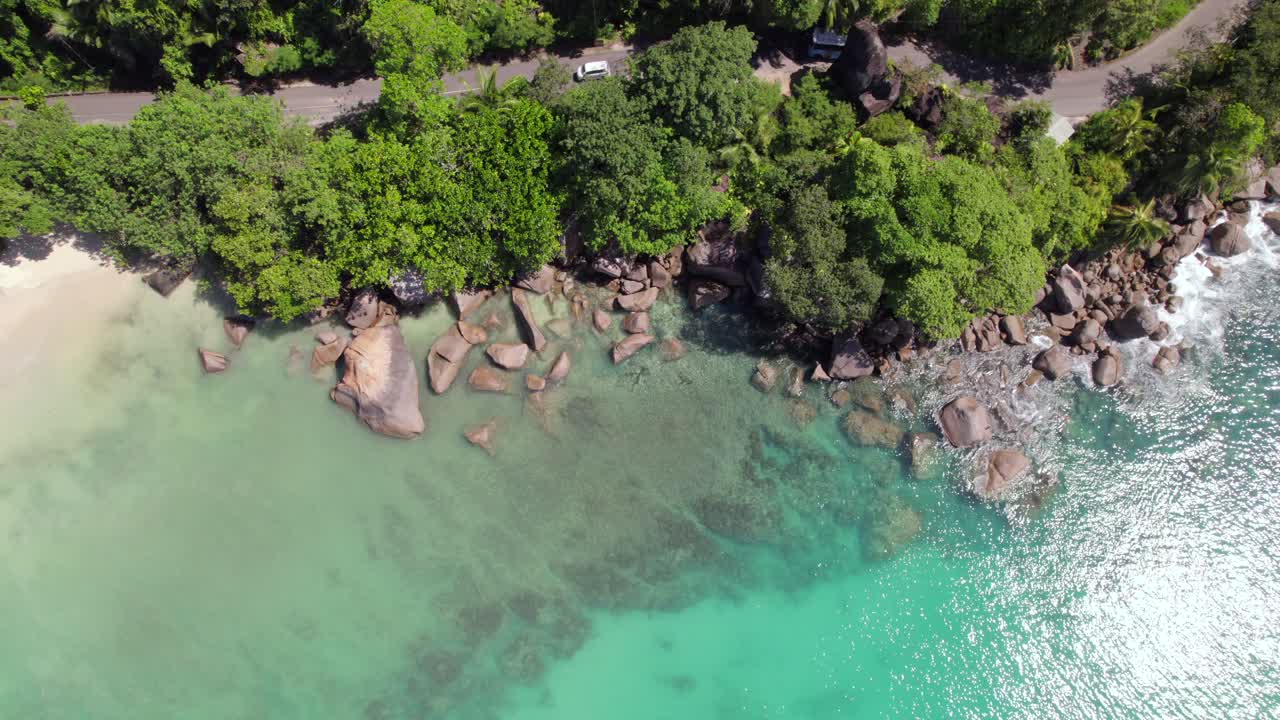 Rekaman drone jalan aspal, kendaraan melintas, batu granit di dekat pantai, dikelilingi pohon, air biru kehijauan, Mahe Seychelles 30 fps 1