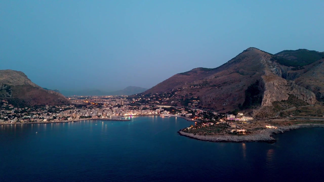 İtalya, Sicilya, Palermo'daki Sferracavallo'da sahil köyünün, aydınlatılmış sokakların ve kayalık sahil şeridinin havadan mavi saat görünümü.