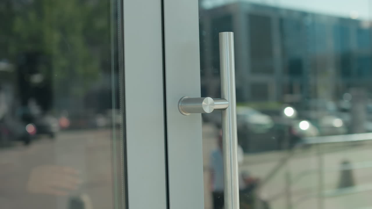 Close up of woman holding bag dressed in black suit walking out of building through glass door with urban street view in background, reflections of people and cars on glass