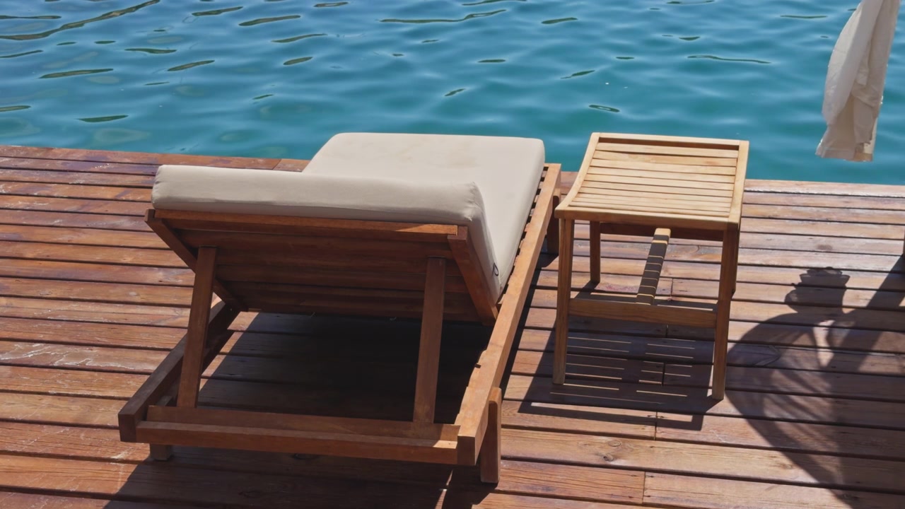 Wide shot of empty sun lounger and side table on sunny wooden dock overlooking bright blue sea, luxury beach club setting, Mediterranean coast, Turkey.