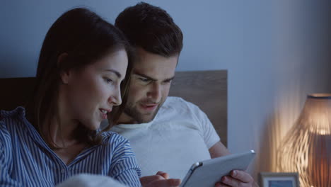 vista de cerca de una joven pareja caucásica sentada en la cama y viendo un video en la tableta por la noche