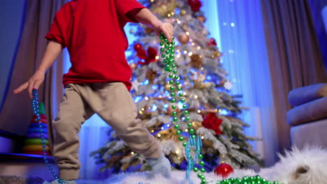 small kid in red cap sits on the floor near the christmas tree. baby boy plays with string of beads. low angle view.