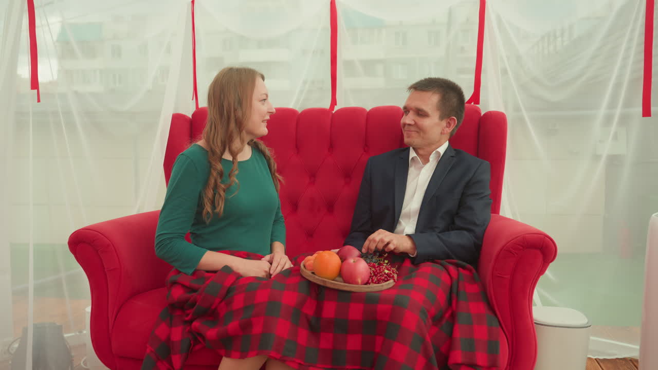 caucasian couple seated on red sofa sharing playful indoor date, woman feeds man pomegranate while both laugh, plaid blanket draped across laps, bowl of fruit at center, bright window with sheer