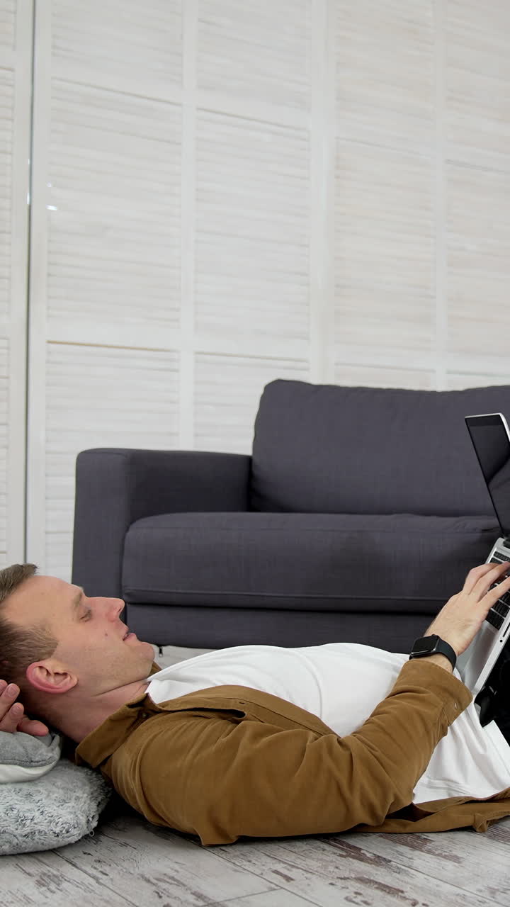 Remote job. Tired freelancer works on a laptop while lying on the floor in the kitchen. Young man studying through the laptop at home. Distance education. Vertical video