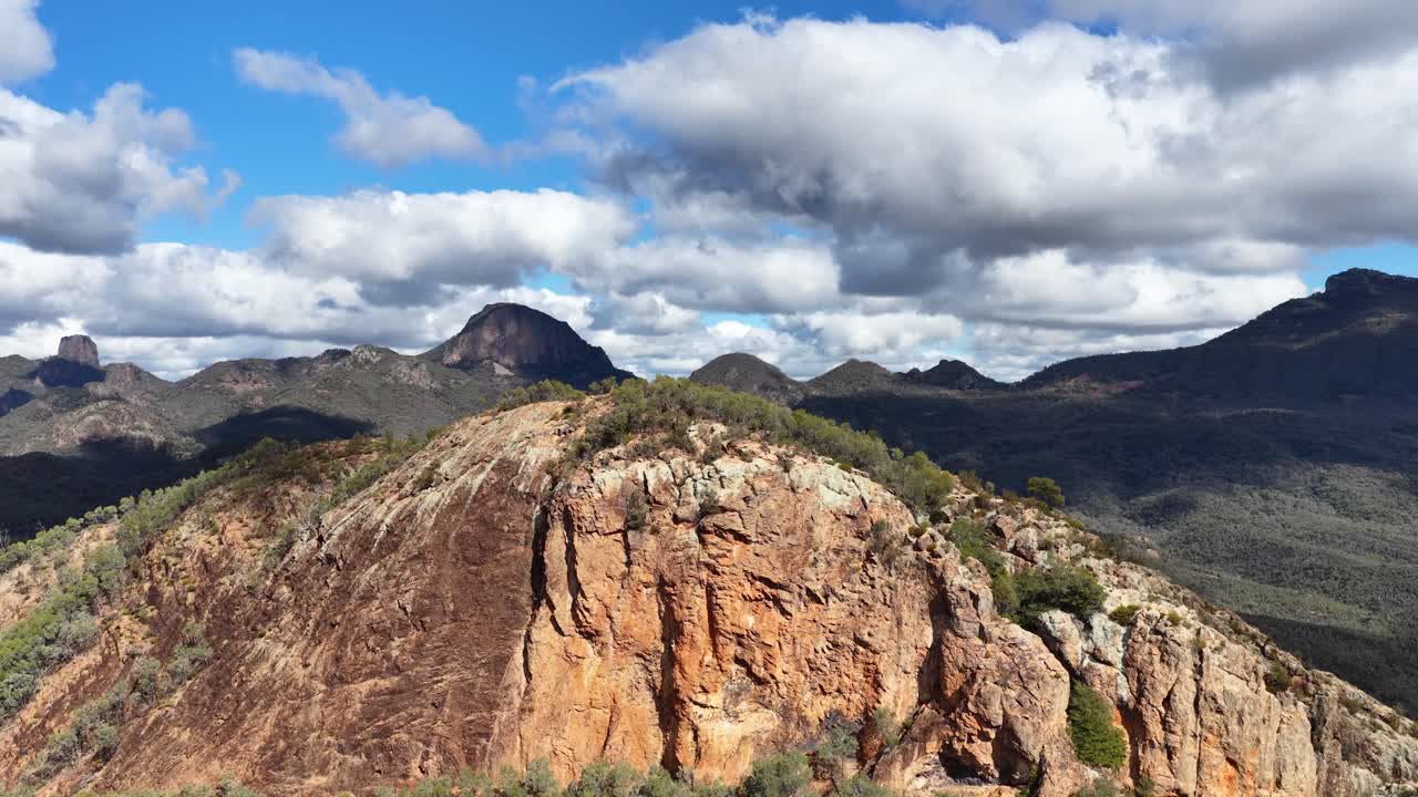 Drone camera glides above Split Rock, revealing rugged sandstone cliffs, green bushland, and distant peaks under bright daylight and scattered clouds
