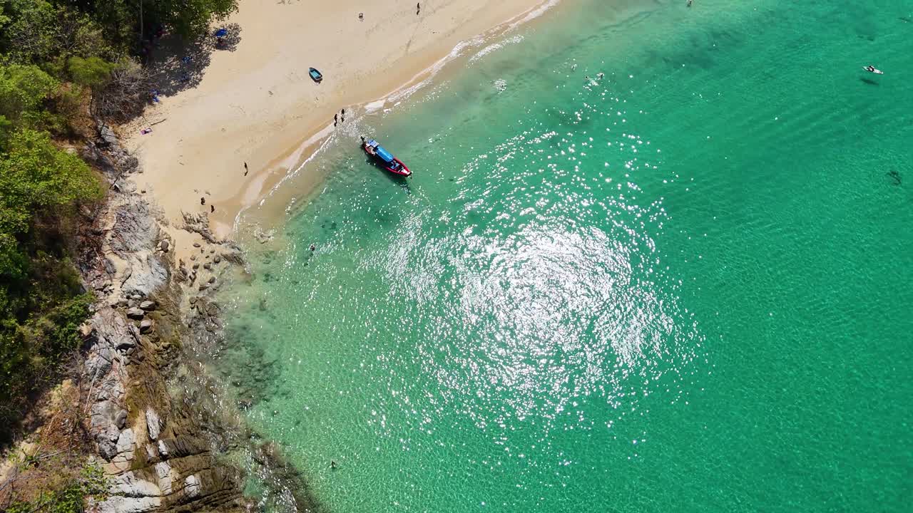 Drone footage captures a vibrant beach in Phuket with turquoise waters, a longtail boat, and tourists enjoying the sun