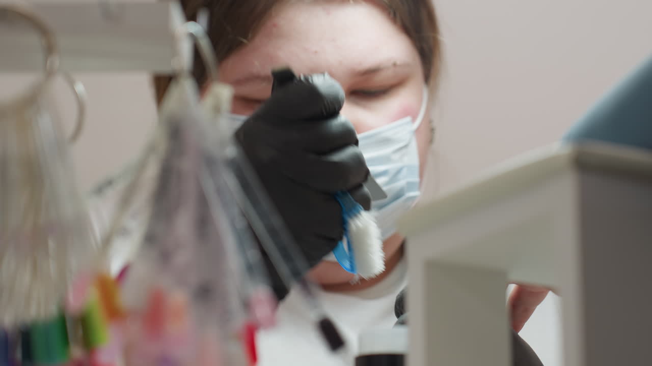 Close up view of nail technician wearing face mask and black gloves filing client nails and brushing off debris while colorful labeled nail polish samples hang in soft blur background