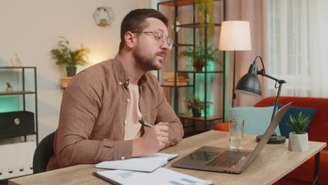 young freelancer writing down notes while attending online meeting on laptop at home office desk