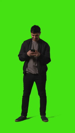 video vertical de larga duración de un joven que envía mensajes de texto en un teléfono móvil de pie contra una pantalla verde