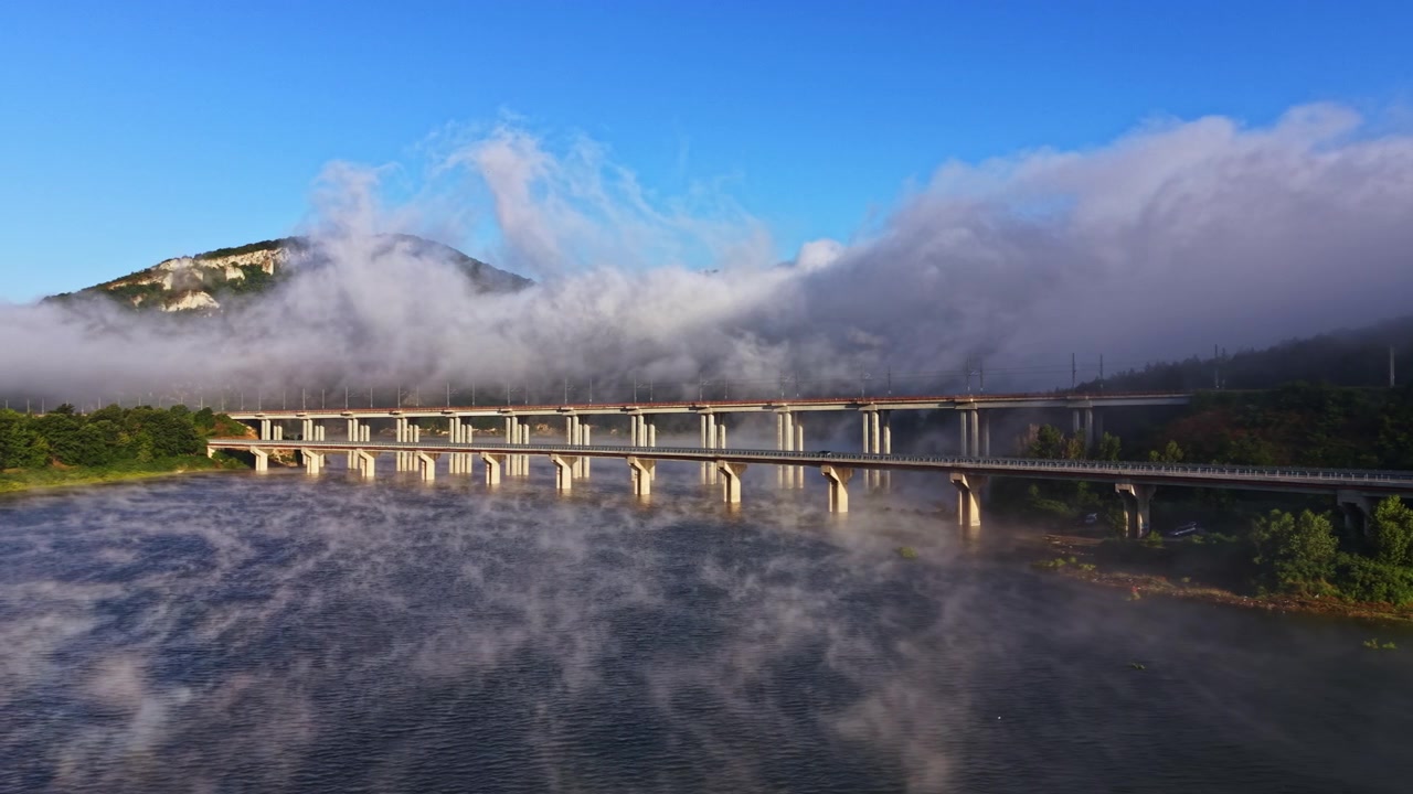 Tåge dækker en bro over en flod med bjerge i baggrunden i de tidlige morgentimer i et naturligt landskab.