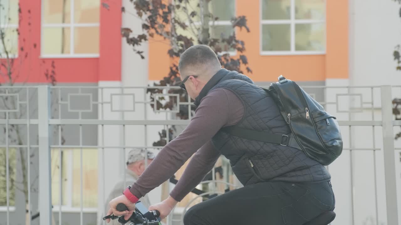 안경과 배을 입고 자전거를 타고 도시를 떠나는 남자의 측면 전망. 산악 자전거에 탑승한 남자가 여름 해가 뜨는 동안 마을의 건물을 타고 간다.